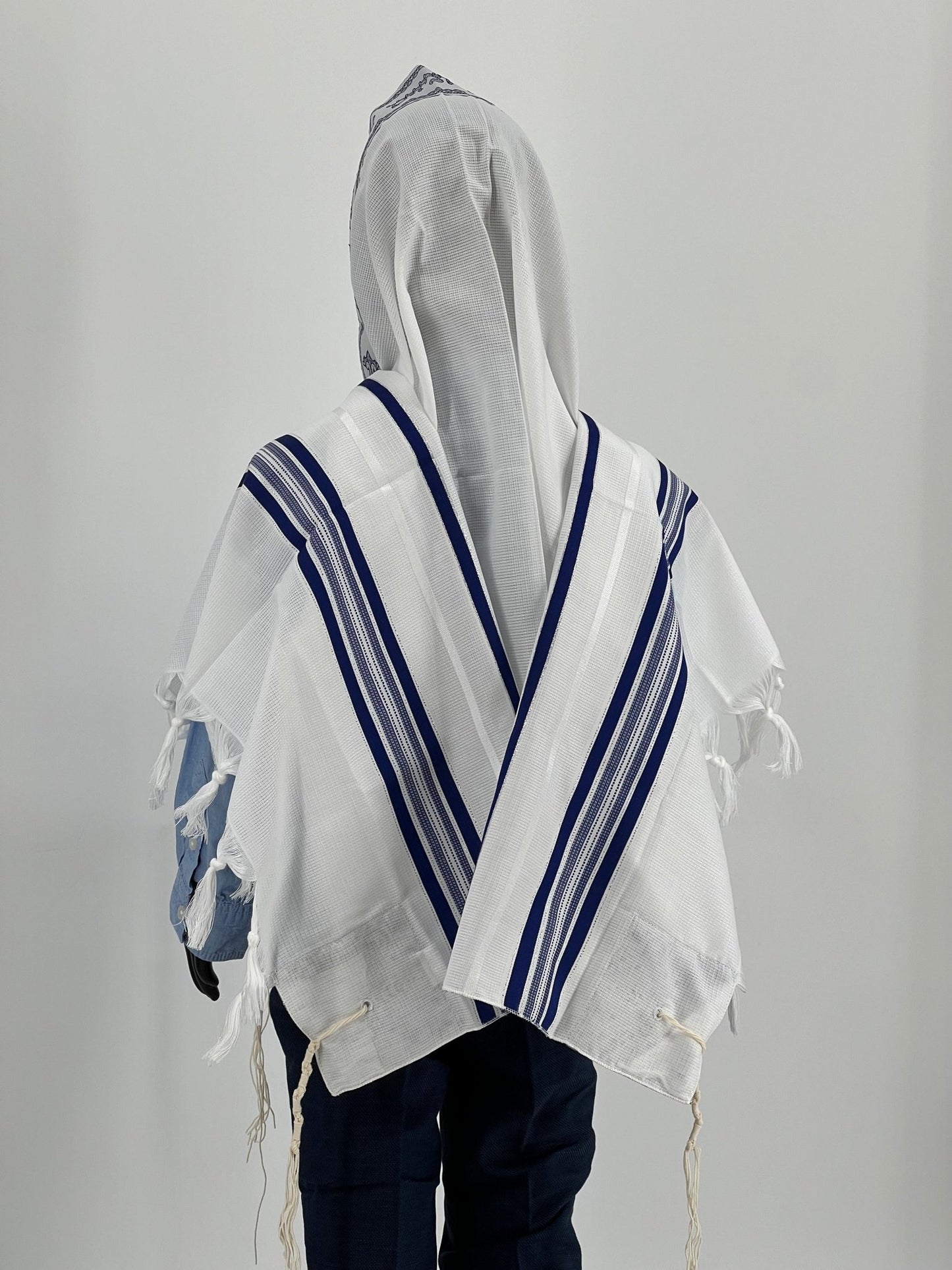 The tallit features traditional tzitzit (fringes) at the corners and is worn over a light blue shirt and dark pants.