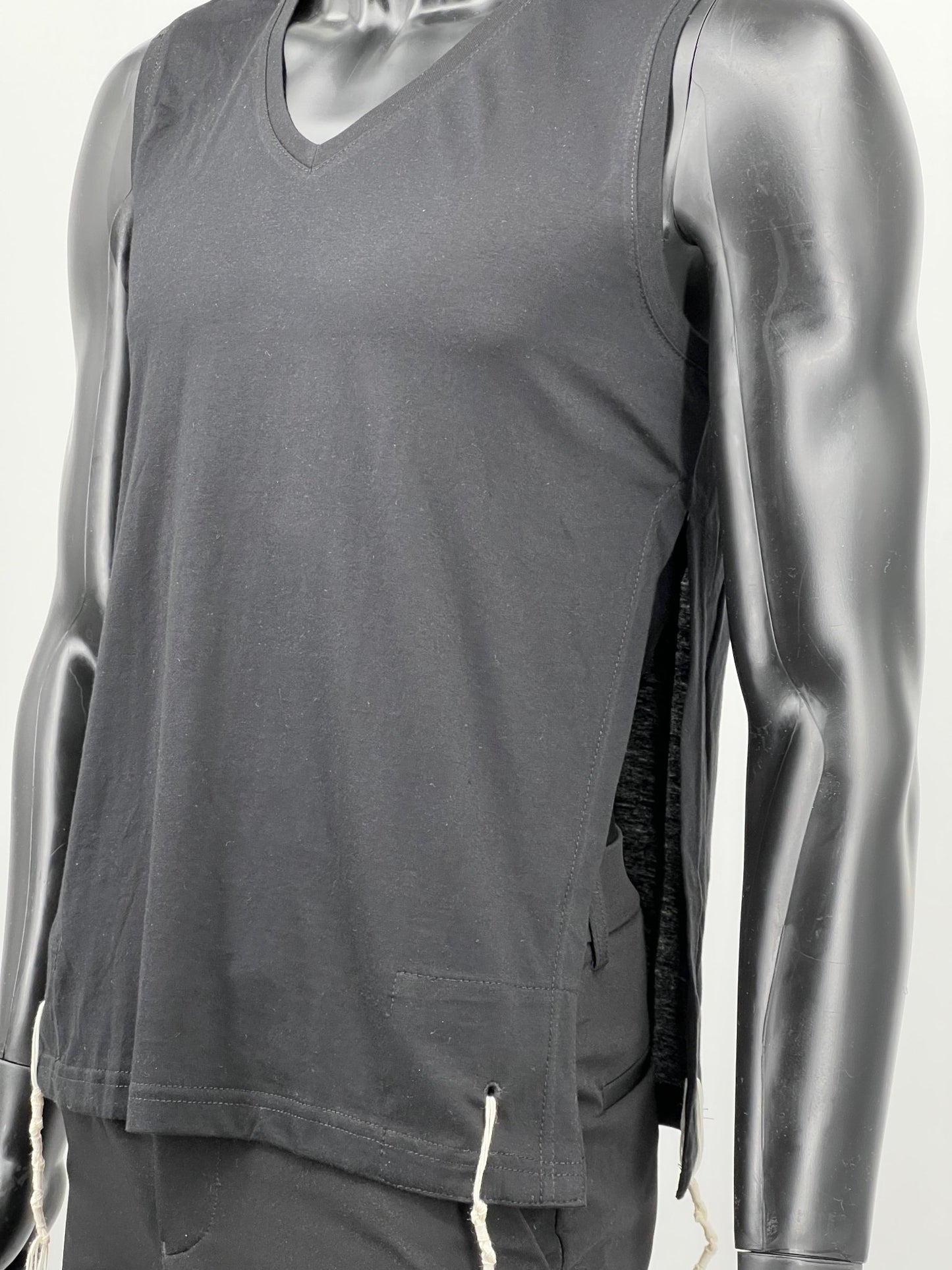 Side angled view of a mannequin wearing a black sleeveless V-neck Tzitzit T-shirt vest. The garment features white Tzitzit fringes hanging from reinforced bottom corners and a side opening detail. It is worn over black pants and displayed against a plain light background.