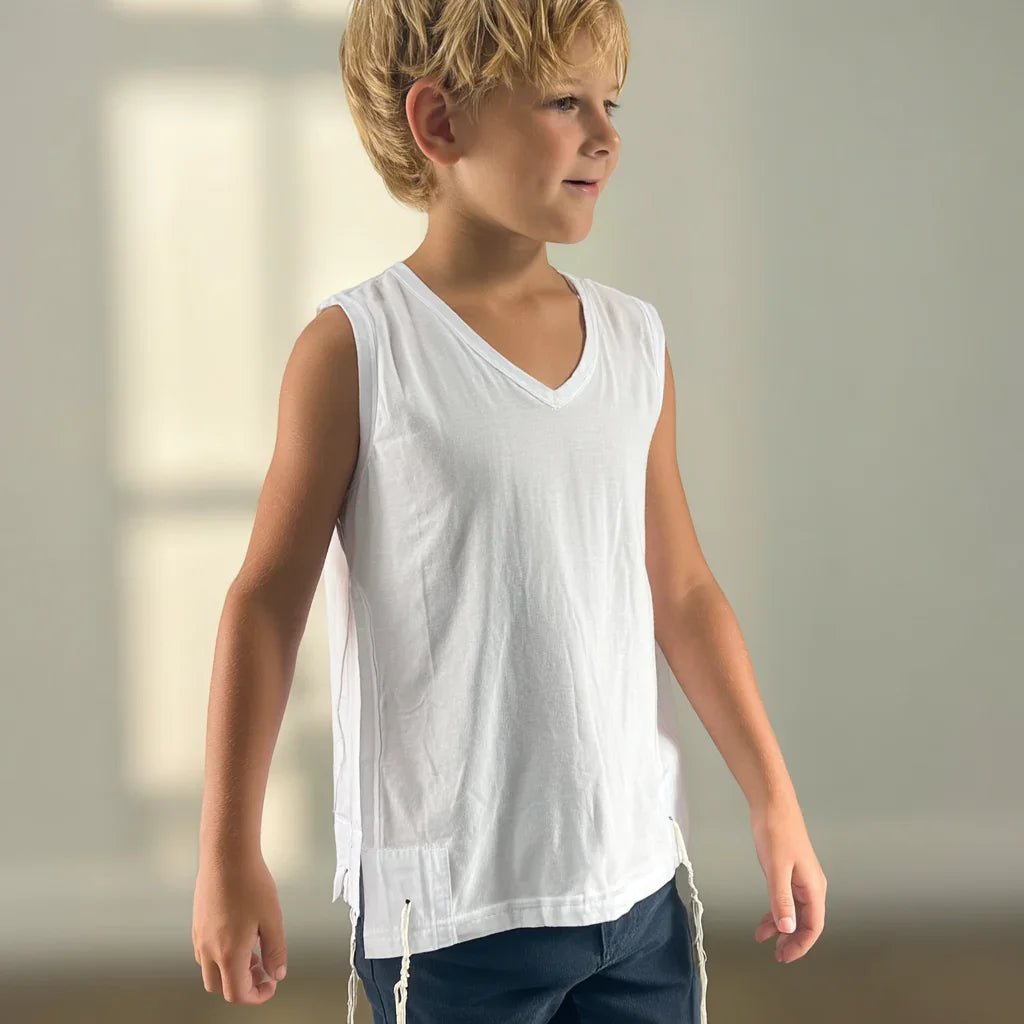 Young boy wearing a white Tzitzit T-shirt sleeveless and dark pants against a neutral background