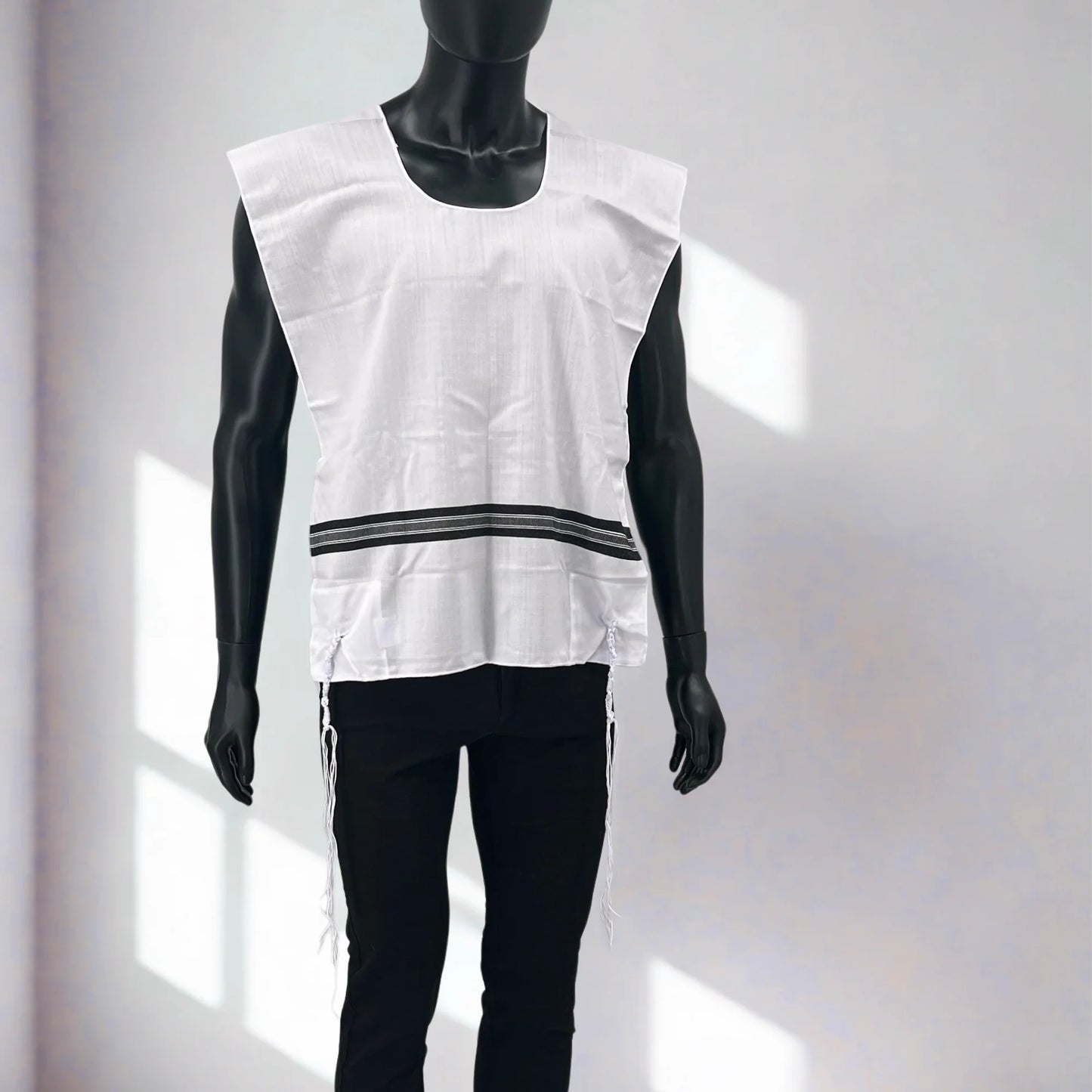 Front view of a black mannequin wearing a white Chabad Tallit Katan with black stripes and a rounded neckline. The garment features long, knotted kosher tzitzit strings hanging from all four corners and is displayed against a softly lit background with window shadows.