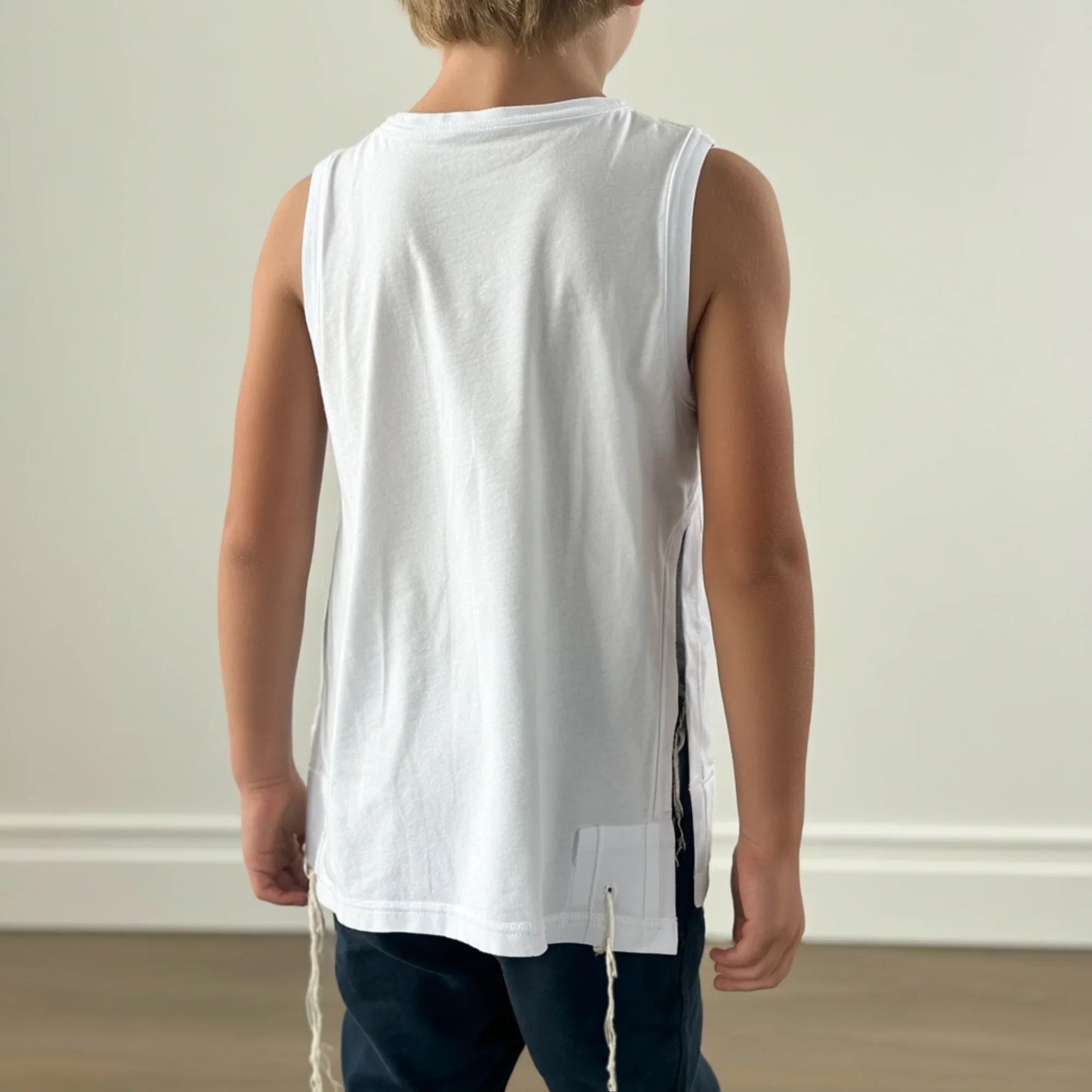 Child wearing a white sleeveless Tzitzit T-shirt, backview, with a subtle pattern on a plain background