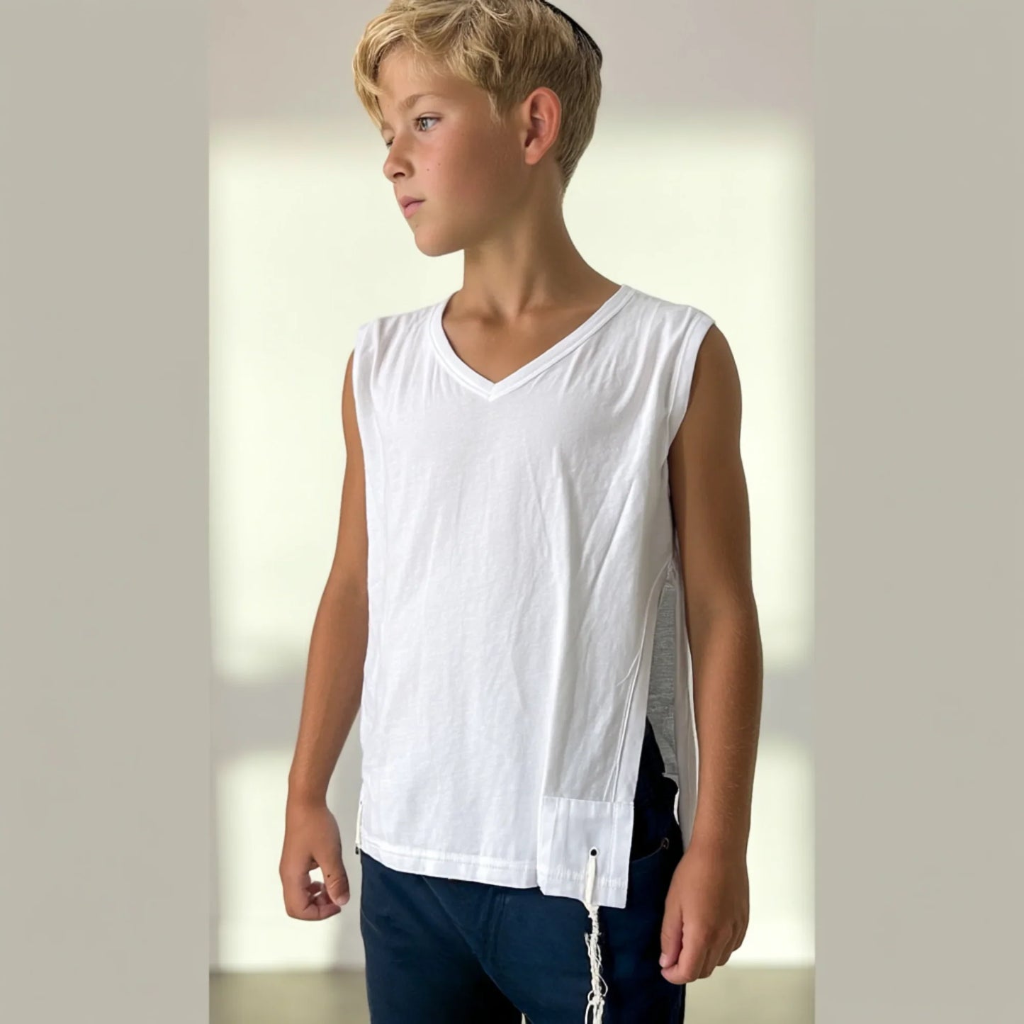 Young boy wearing a white sleeveless Tzitzit T-shirt and dark pants against a plain background