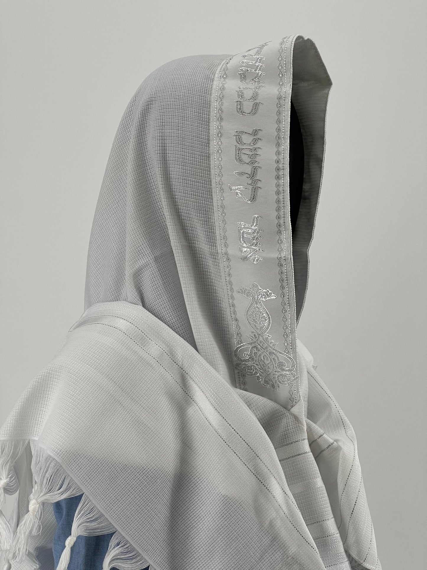 Child mannequin wearing a Jewish prayer shawl with an embroidered silver Hebrew blessing along the edge of the hood.