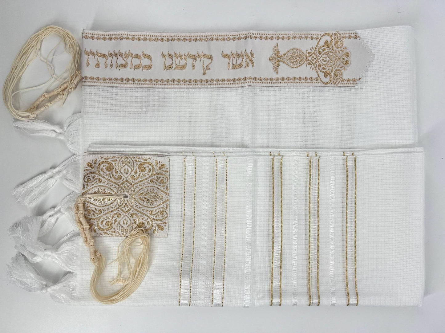 Folded white tallit prayer shawl with decorative gold embroidery, featuring tzitzit fringes and an ornate atarah