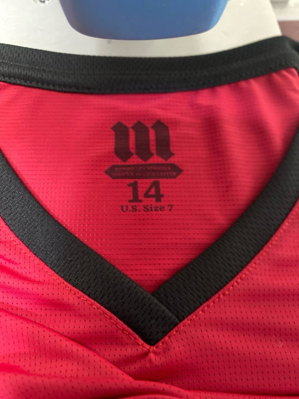 Close-up image of the inside collar of a red athletic Tzitzit T-shirt with a black V-neck trim. The label printed on the fabric includes the MMJ Judaica logo