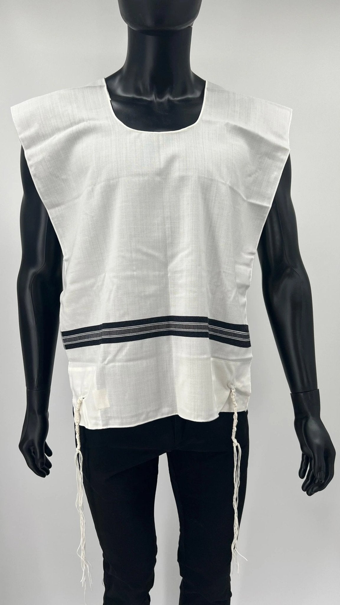Child-sized mannequin wearing a white wool Tallit Katan with black striping and a rounded neckline. The garment includes kosher tzitzit strings hanging from all four corners and is designed according to traditional Jewish standards.