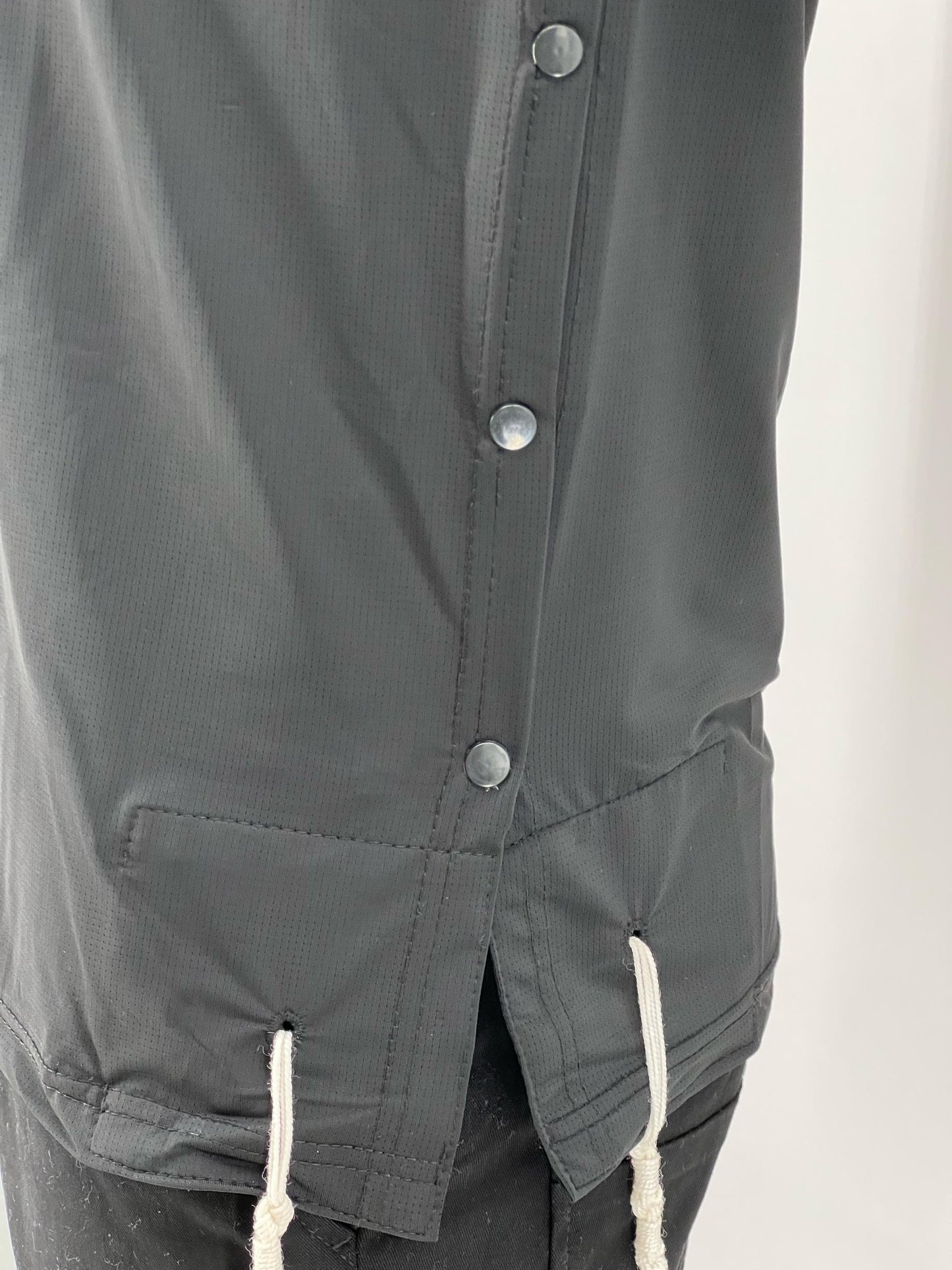 Close-up of the side view of a black moisture-wicking Tzitzit T-shirt, highlighting the snap buttons, side slit, reinforced stitching, and two sets of white Tzitzit strings hanging from the corners.