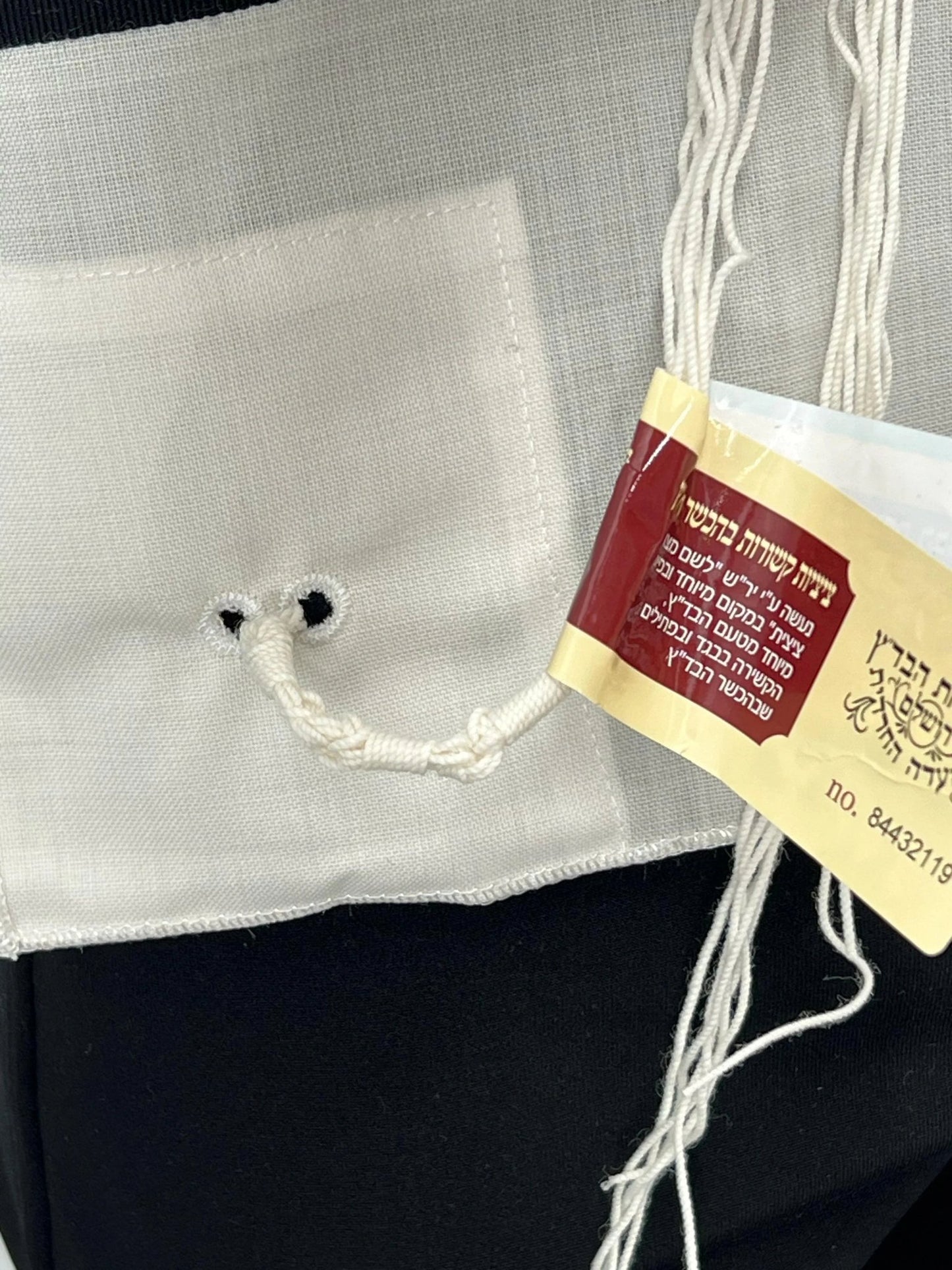 Close-up of the bottom corner of a white Tallit Katan garment, showing a pair of reinforced holes with a looped tzitzit string tied through them. A red and yellow kosher certification tag in Hebrew is attached, indicating the tzitzit were handmade according to halachic standards.