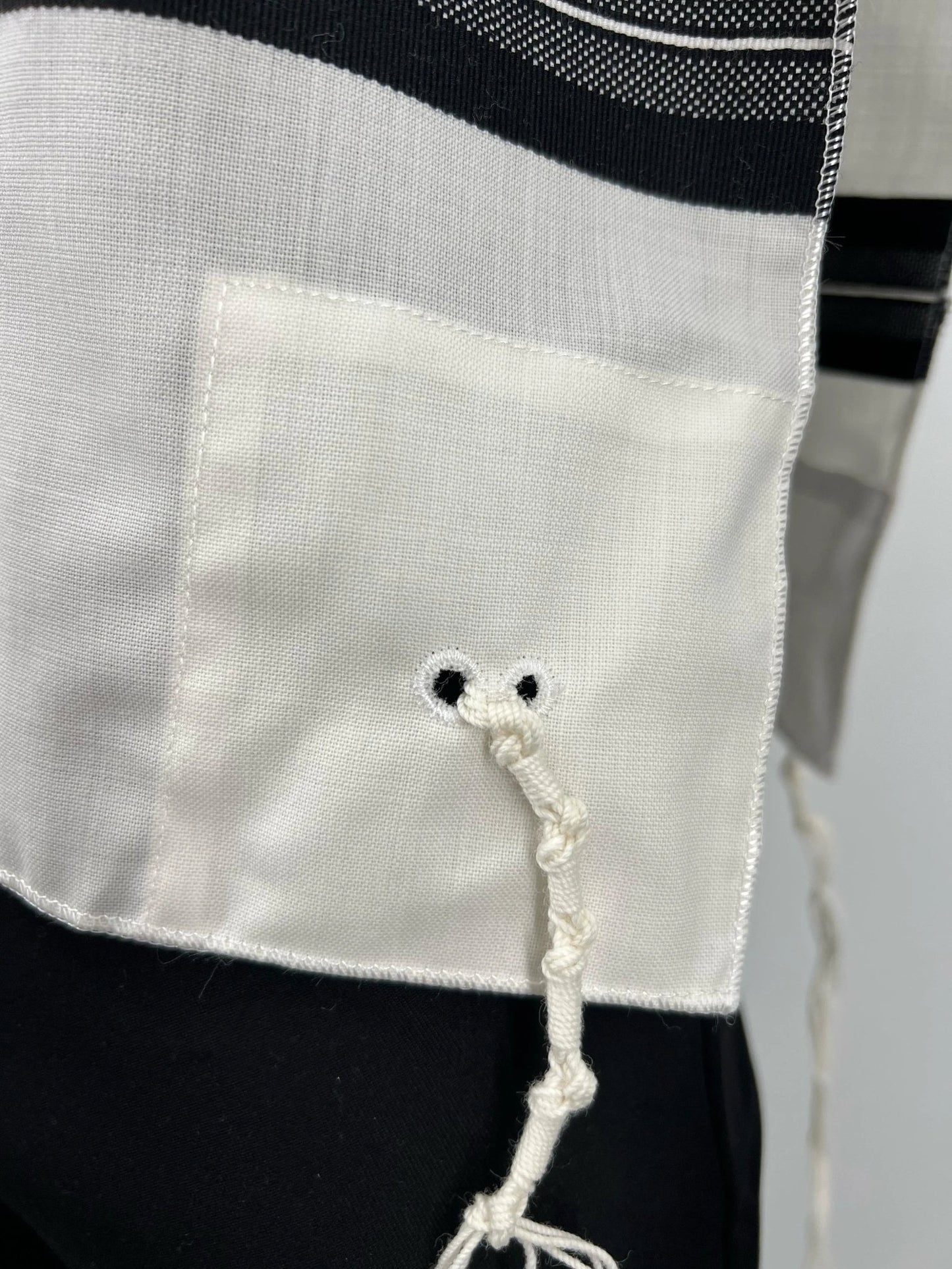 Close-up of a corner of a white wool Tallit Katan featuring black striping and a reinforced fabric patch with two black-stitched holes. A kosher tzitzit string is threaded and tied through the holes in accordance with halachic standards.