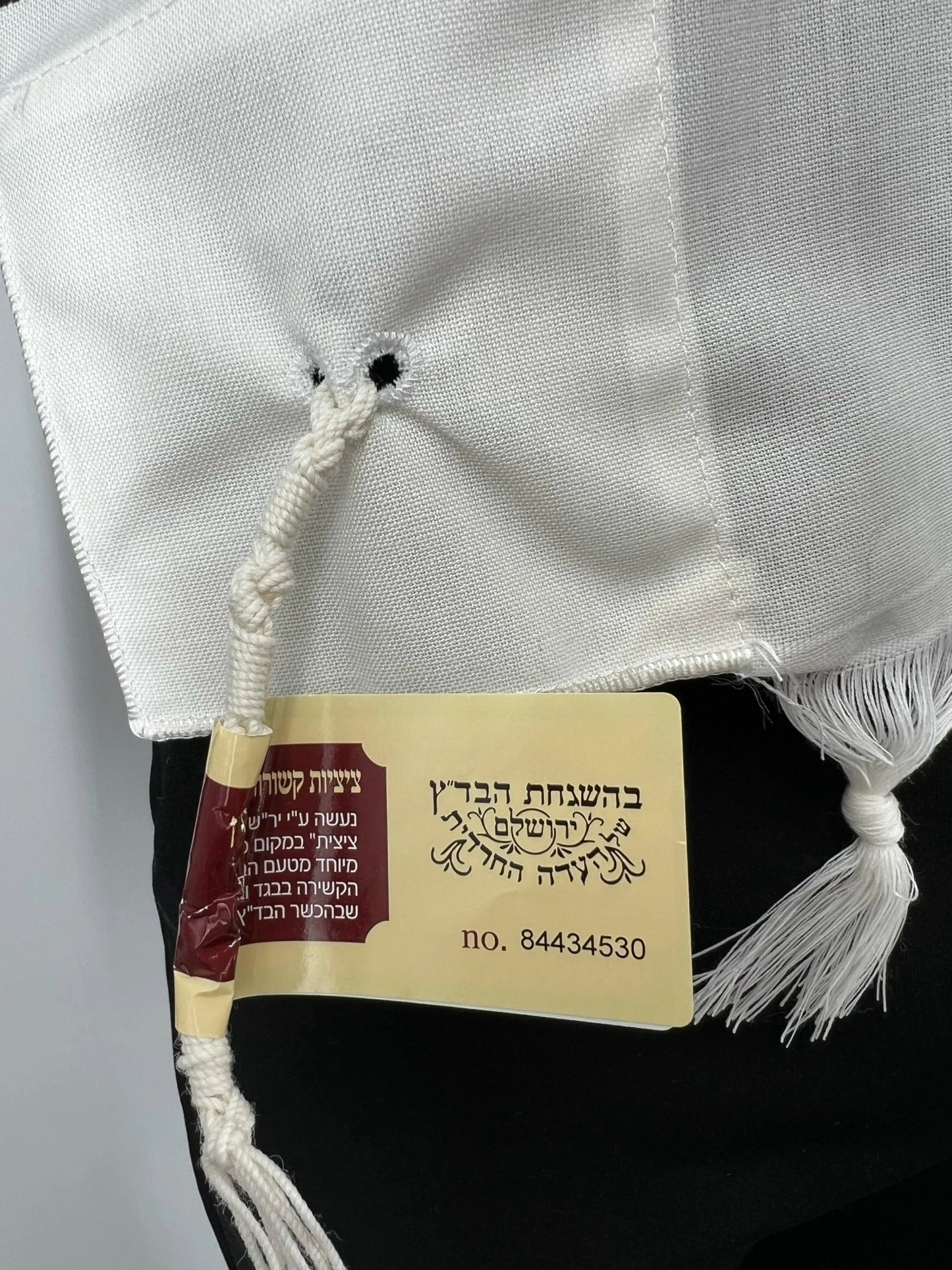 red and yellow kosher certification tag in Hebrew is attached, indicating the tzitzit were handmade according to halachic standards.