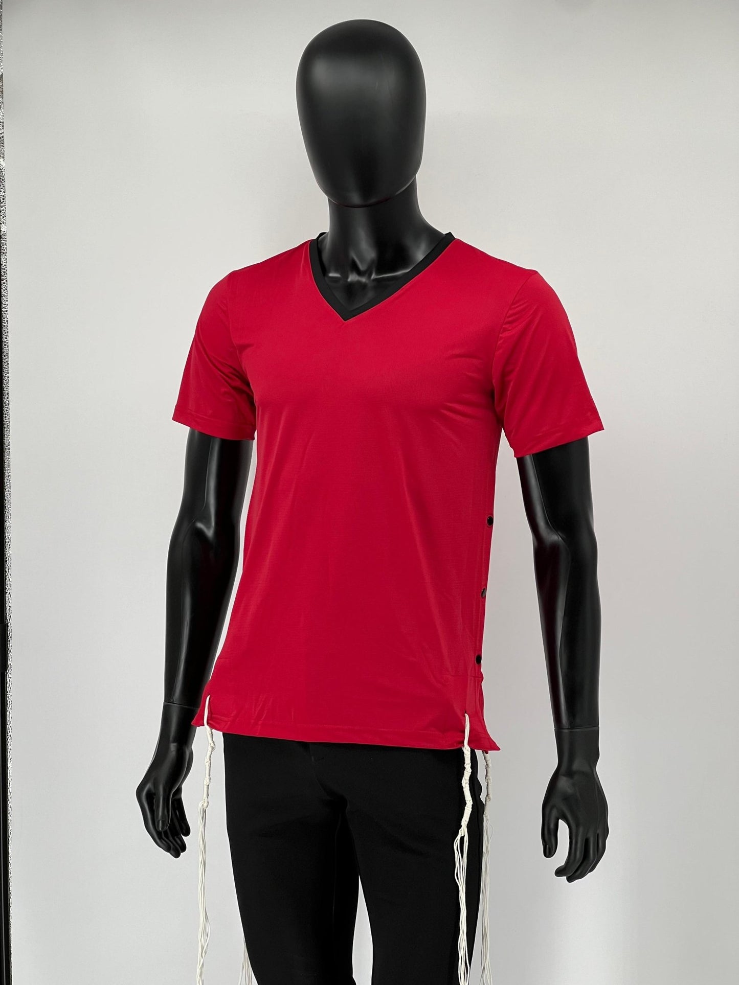 Front view of a red athletic Tzitzit T-shirt with a black V-neck, worn by a black mannequin. The shirt features side snap closures and traditional white Tzitzit strings hanging from the hem, combining halachic requirements with a modern sporty design.