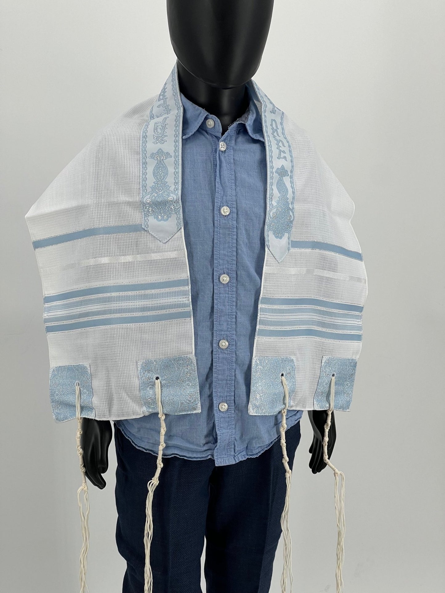 Child mannequin wearing a white children's tallit (Jewish prayer shawl) with light blue stripes and embroidered corners