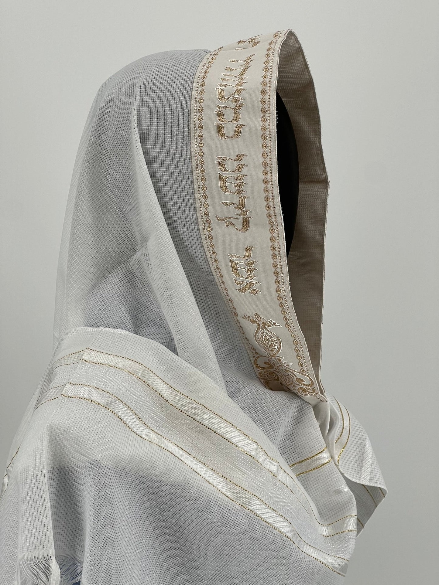 Child mannequin wearing a white tallit with an embroidered gold Hebrew blessing along the edge of the hood.
