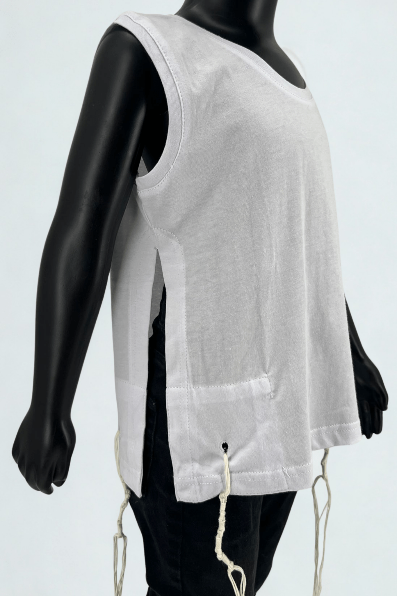 Close-up side view of a child mannequin wearing a sleeveless white Tzitzit T-shirt with halachically tied fringes (Tzitzit) on the corners. The shirt features wide side openings and reinforced stitching, shown paired with black pants, against a light background.