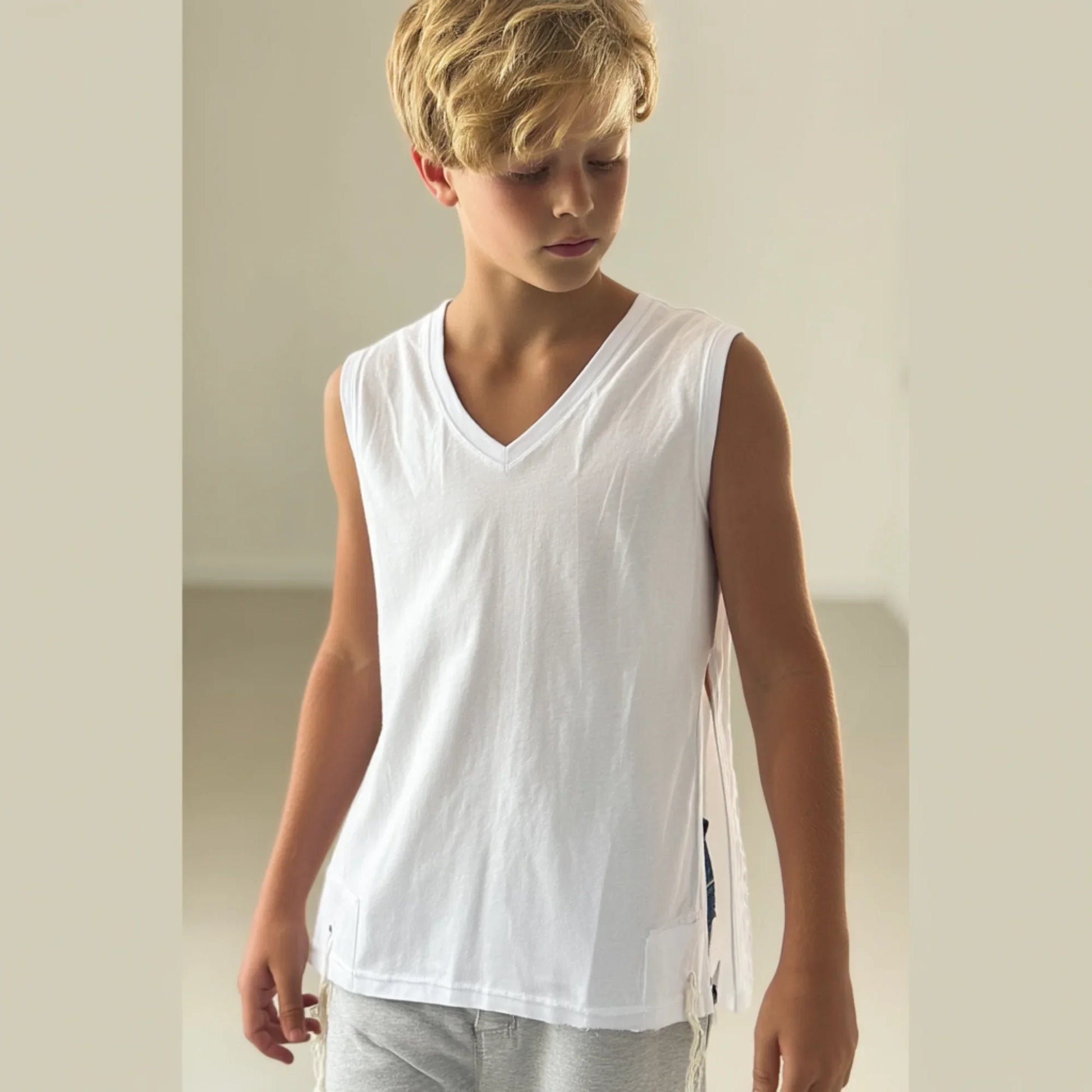 Young boy wearing a white Tzitzit T-shirt sleeveless and gray pants against a plain background
