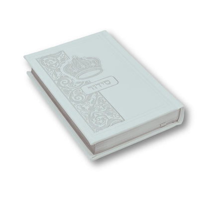 White faux leather Siddur with an embossed crown and decorative floral design on the cover. The Hebrew word "סידור" appears in silver foil in the center. The book is slightly angled and closed, showing its elegant edges and craftsmanship.
