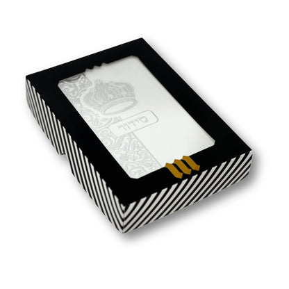 White faux leather Siddur with silver embossed crown design, displayed in a black-and-white striped gift box with a clear window. Hebrew word "סידור" appears on the cover. The box features a gold "MMJ" logo at the bottom.