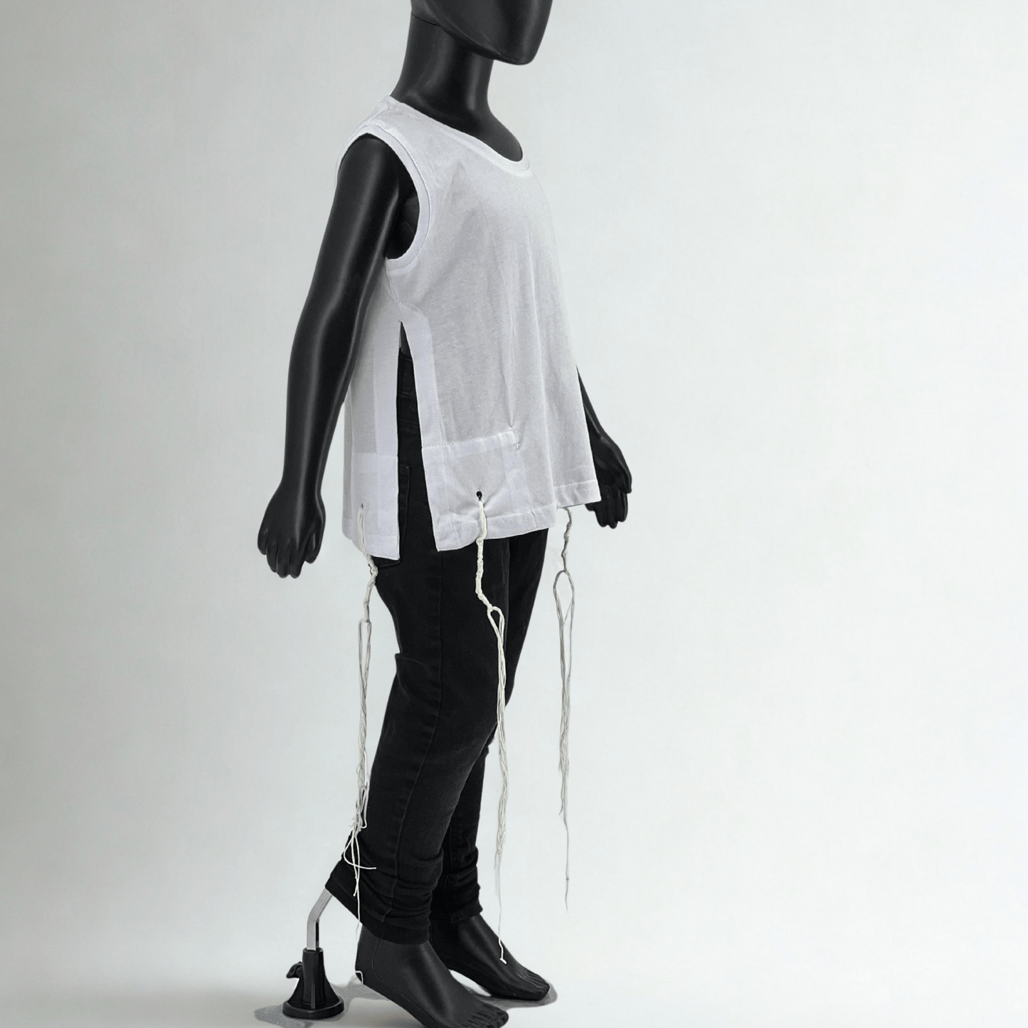 Child-sized mannequin wearing a sleeveless white Tzitzit T-shirt with side openings and tied fringes (Tzitzit) at all four corners, paired with black jeans, photographed in a studio with a plain white background.