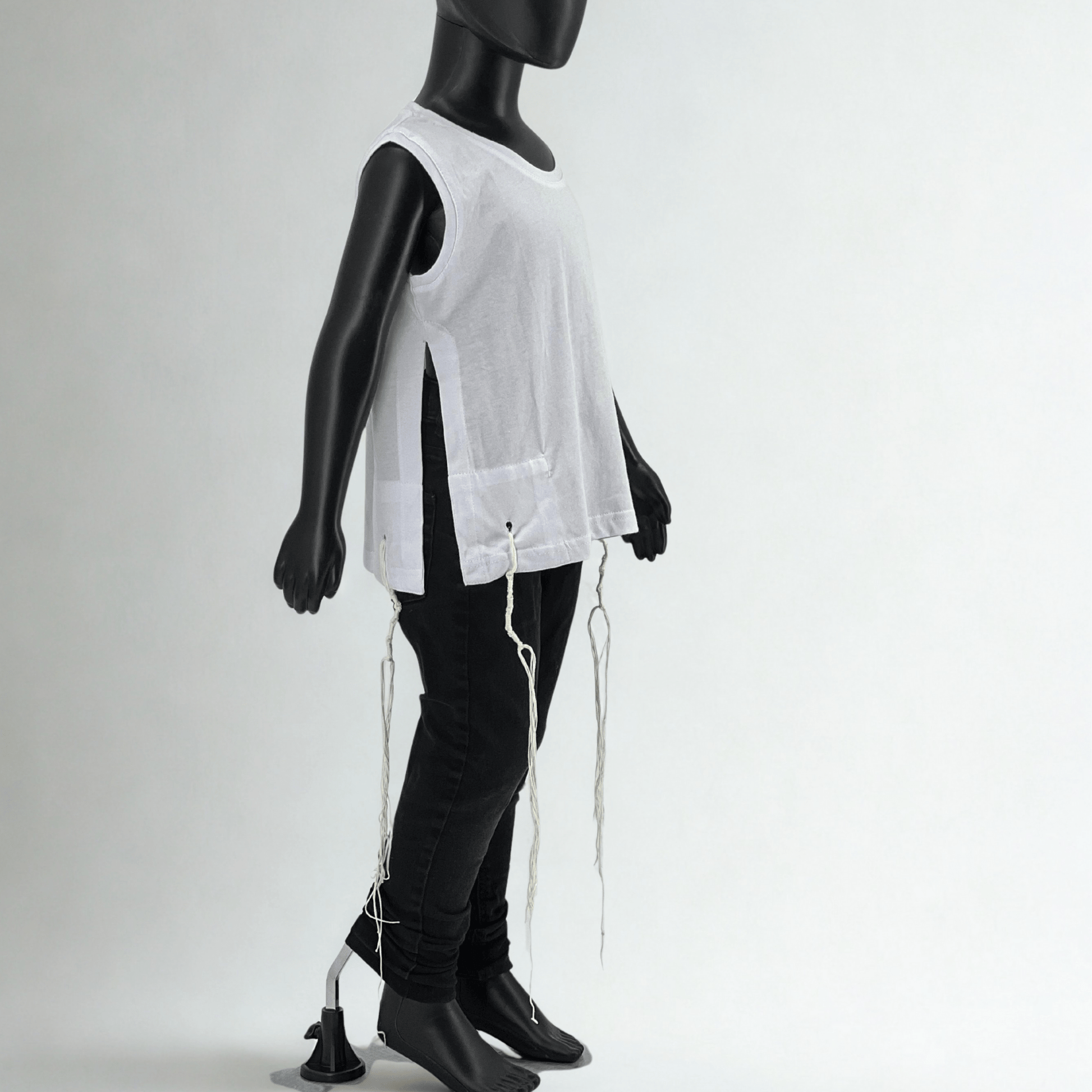 Child-sized mannequin wearing a sleeveless white Tzitzit T-shirt with side openings and tied fringes (Tzitzit) at all four corners, paired with black jeans, photographed in a studio with a plain white background.