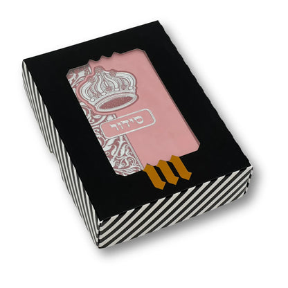 Pink faux leather Siddur with silver embossed crown design, displayed in a black-and-white striped gift box with a clear window. Hebrew word "סידור" appears on the cover. The box features a gold "MMJ" logo at the bottom.