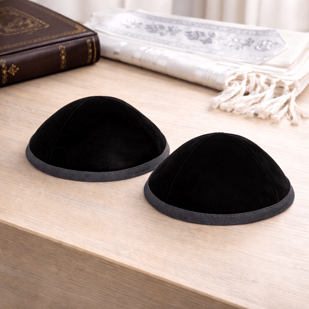 Two black kippah on a wooden surface with books and a decorative item in the background.
