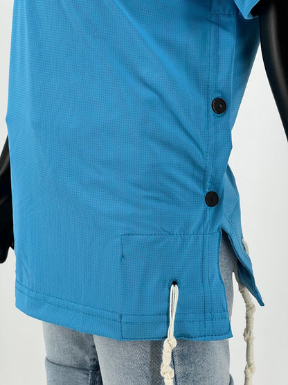 Side view of a bright blue Tzitzit T-shirt worn over light blue denim shorts. The shirt features two visible black buttons, a side slit, and two white Tzitzit fringes hanging from reinforced holes at the hem. A mannequin arm is partially visible on the right side.