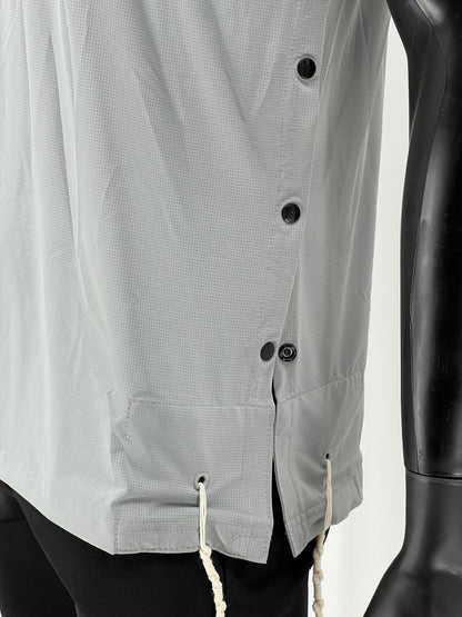 Close-up side view of a light gray Tzitzit T-shirt featuring four black buttons along a side opening and two white Tzitzit fringes hanging from reinforced holes near the hem. The shirt is made from breathable mesh fabric and worn over black pants. A mannequin arm is visible on the right side.