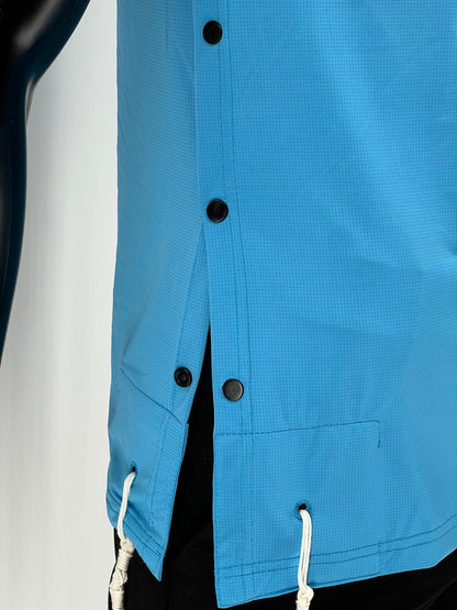 Close-up side view of a bright blue Tzitzit T-shirt showing four black side buttons and two white Tzitzit fringes hanging from reinforced corner holes. The breathable fabric and precise stitching are clearly visible. The garment is worn over black pants and displayed against a plain light background
