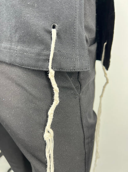 Close-up view of the bottom corner of a black Tzitzit T-shirt vest, showing a white Tzitzit fringe hanging from a reinforced hole. The garment is worn over black pants, and the detailed stitching of the hem and fabric texture are clearly visible.