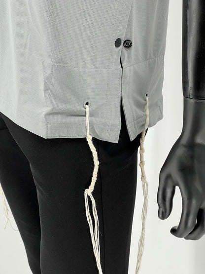 Close-up view of the lower side of a light gray Tzitzit T-shirt, highlighting two black side buttons, a small side slit, and two white Tzitzit fringes hanging from reinforced corner holes. The shirt is made from breathable fabric and worn over black pants. A mannequin hand is visible on the right side of the image.