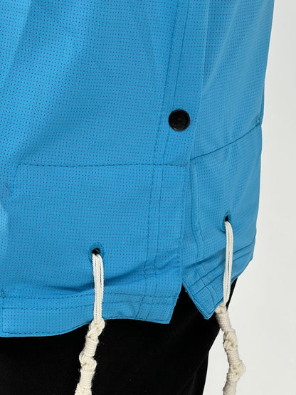 Close-up of the lower side of a bright blue Tzitzit T-shirt featuring a black side button and two white Tzitzit fringes hanging from reinforced holes. The breathable mesh fabric and detailed stitching are clearly visible. The shirt is worn over black pants and displayed against a plain light background.