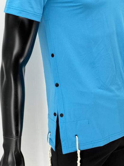 Side view of a bright blue Tzitzit T-shirt on a mannequin, showing four black side buttons and two white Tzitzit fringes hanging from reinforced corner holes. The shirt is made from breathable, perforated fabric and is paired with black pants. Displayed against a plain light background.