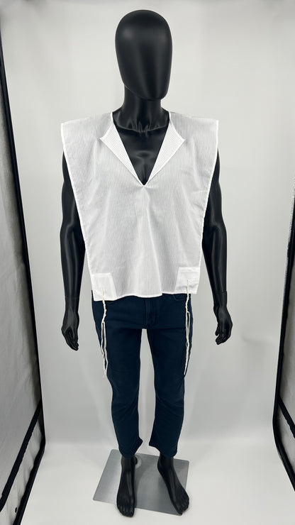 A full-body view of a black mannequin wearing a white cotton Tallit Katan with subtle vertical stripes. The garment has a V-neck opening and features long kosher tzitzit strings hanging from all four corners. The mannequin is dressed in dark pants, and the tallit katan is worn over them.