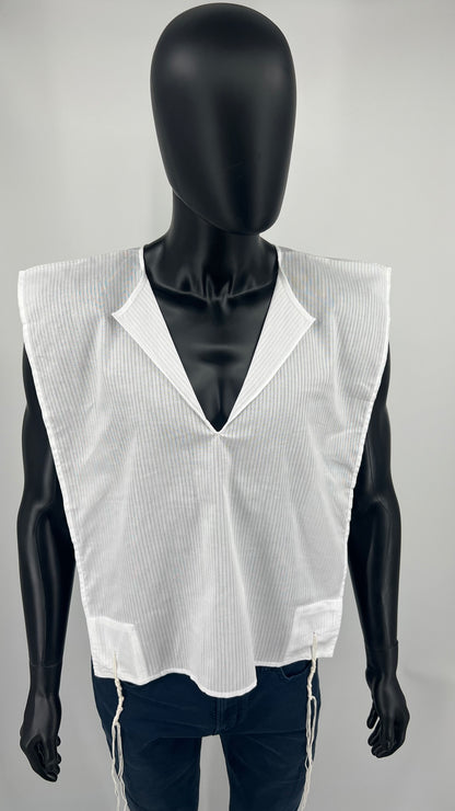 A full-body view of a black mannequin wearing a white cotton Tallit Katan with subtle vertical stripes. The garment has a V-neck opening and features long kosher tzitzit strings hanging from all four corners. The mannequin is dressed in dark pants, and the tallit katan is worn over them.