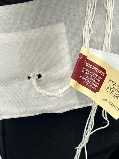 Close-up of the bottom corner of a white Tallit Katan garment, showing a pair of reinforced holes with a looped tzitzit string tied through them. A red and yellow kosher certification tag in Hebrew is attached, indicating the tzitzit were handmade according to halachic standards.