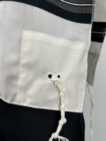 Close-up of a corner of a white wool Tallit Katan featuring black striping and a reinforced fabric patch with two black-stitched holes. A kosher tzitzit string is threaded and tied through the holes in accordance with halachic standards.