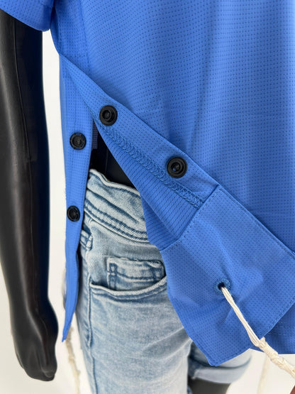 Close-up of the side view of a blue Tzitzit T-shirt showing three black buttons along the side opening. The shirt is worn over light blue denim shorts, and one white Tzitzit fringe is visible hanging from a corner hole. The breathable, mesh fabric texture and reinforced stitching are clearly shown.