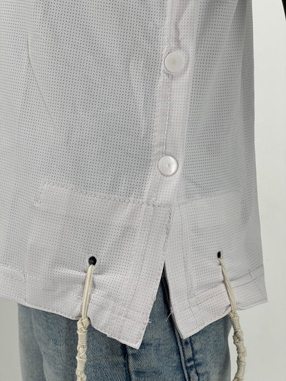 Close-up view of the side of a white perforated Tzitzit T-shirt featuring two white side buttons and a small side slit at the hem. Two white Tzitzit fringes are visible hanging from reinforced holes at the garment's corner. The shirt is worn over light blue denim shorts.
