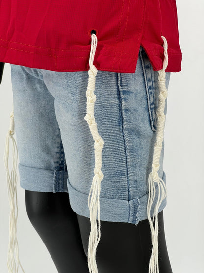 Close-up view of the lower side of a red Tzitzit T-shirt worn over light blue denim shorts. Two white Tzitzit fringes are visible hanging from reinforced corner holes near a side slit in the hem. The image highlights the detailed stitching and fabric texture.