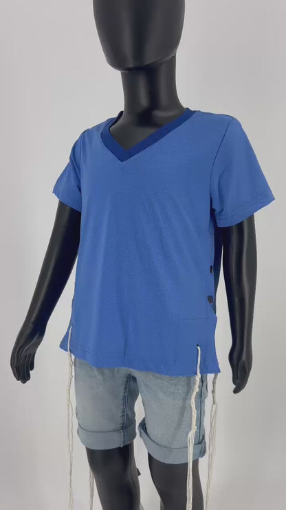 Video of Mannequin dressed in a blue V-neck short-sleeve Tzitzit T-shirt with a breathable mesh texture, featuring white Tzitzit fringes hanging from the garment's corners. The shirt includes white side buttons and is paired with light blue denim shorts. Displayed against a plain light background.