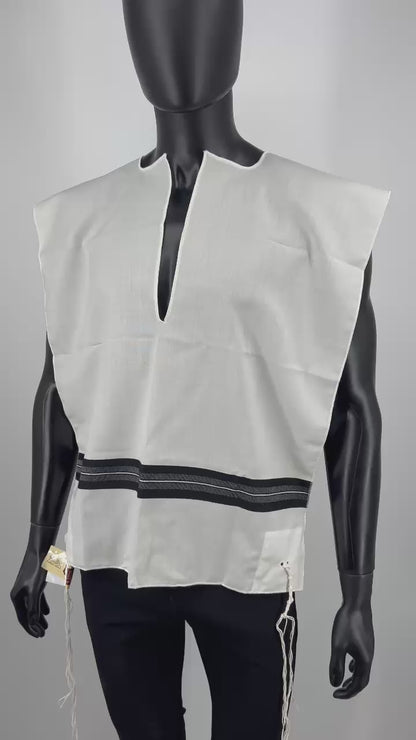 Video of Front view of a black mannequin wearing a white wool Tallit Katan with black striping and decorative fringe along the bottom. The garment features a halachic V-neck opening and long, knotted kosher tzitzit strings hanging from all four corners.